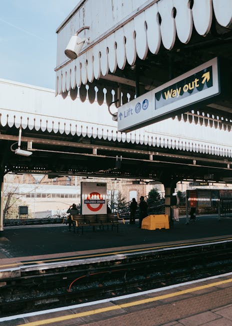 An exterior view of South Wimbledon Station featuring a modern multi-level staircase with metal railings and steps, adjacent to railway tracks. The station's entrance is illuminated by soft lighting, 